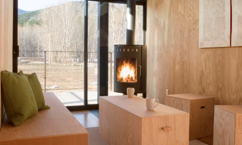 Cozy interior with wooden furniture, a fireplace, and large windows showcasing a natural landscape outside.