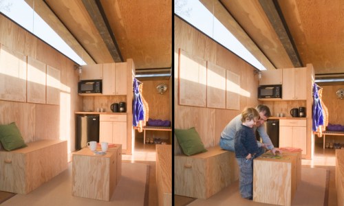 A cozy wooden interior of a small cabin, featuring a man and child playing at a table with kitchen appliances in the background.