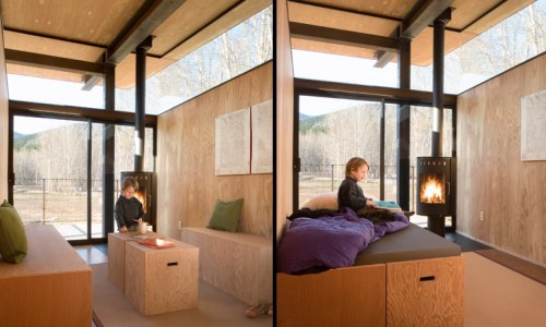 A cozy interior of a modern cabin with a child sitting by a fireplace, large windows, and wooden walls.