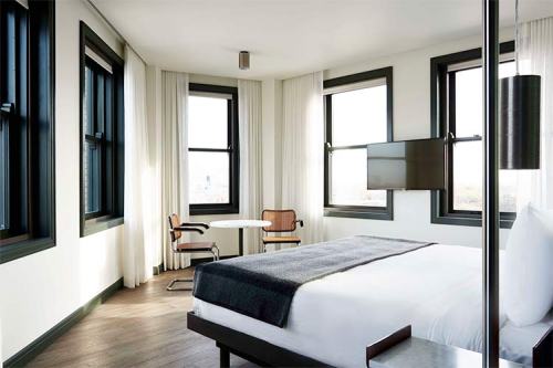 Modern hotel room featuring a bed, a small dining table, large windows, and a wall-mounted TV. Bright and airy atmosphere.
