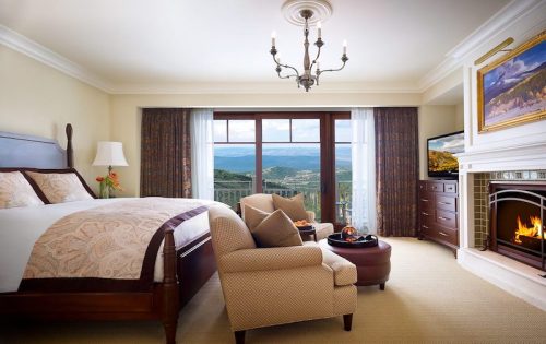 Luxurious hotel room with a king bed, seating area, fireplace, and large windows showcasing a scenic mountain view.