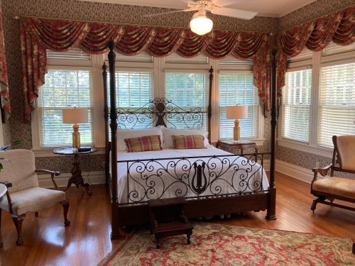 A cozy bedroom featuring a four-poster bed, patterned curtains, and large windows with natural light.