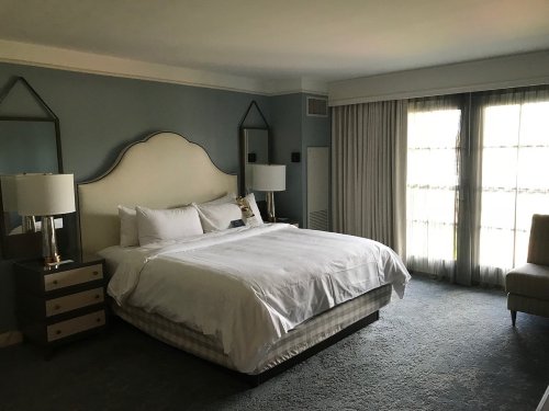 A spacious hotel room featuring a large bed, two lamps, a chair, and large windows with sheer curtains.