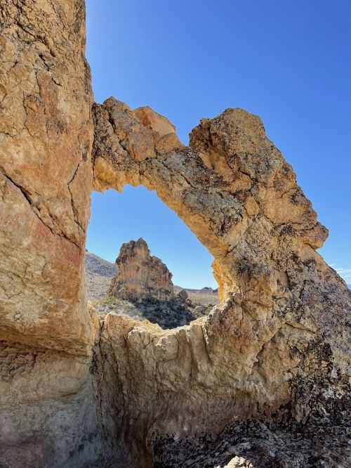 A natural rock formation with a large arch, framed by rugged terrain and a clear blue sky in the background.