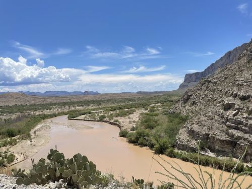 A winding river flows through a desert landscape, surrounded by rocky cliffs and sparse vegetation under a blue sky.
