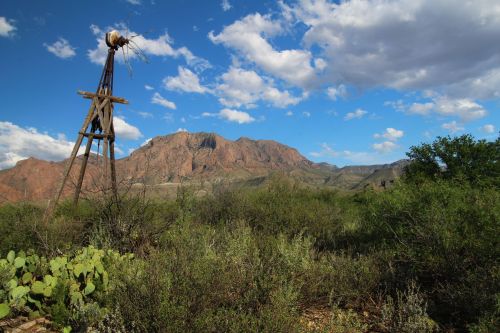 A windmill stands in a desert landscape with mountains and blue skies in the background, surrounded by greenery.