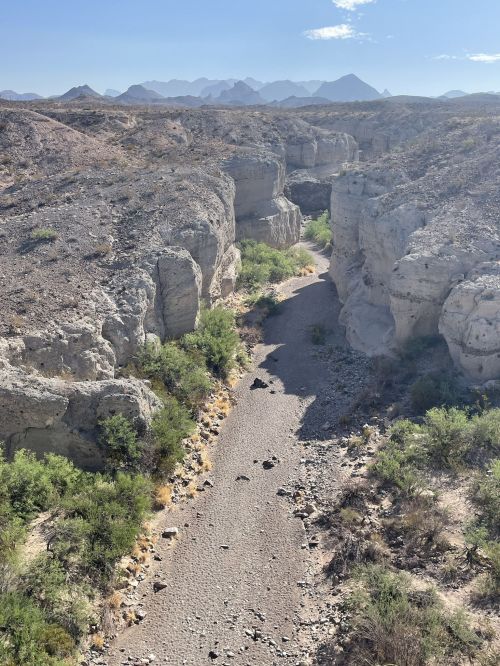 A dry riverbed winds through rocky canyons, surrounded by sparse vegetation and distant mountains under a clear sky.
