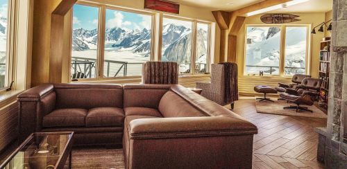 Cozy living room with a brown sofa, large windows showcasing snowy mountains and a glacier view.