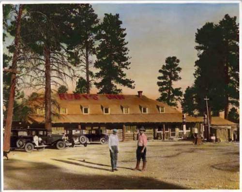 Historic image of Ruby's Inn with vintage cars and two men walking in front, surrounded by tall trees.
