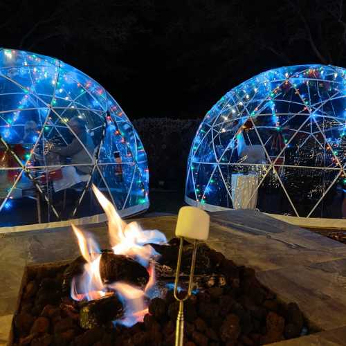 Two illuminated igloo-shaped structures with a fire pit in the foreground, creating a cozy outdoor atmosphere.