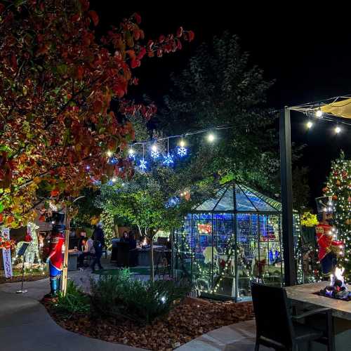 A festive outdoor scene with colorful lights, a decorated tree, and a glass greenhouse surrounded by autumn foliage.