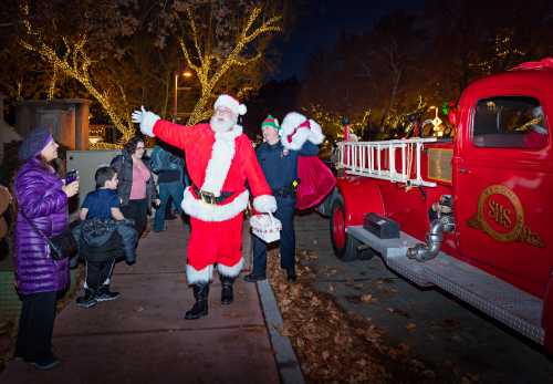 Santa Claus waves to a crowd near a vintage fire truck, surrounded by festive lights and holiday decorations.