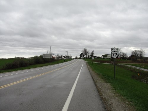 A rural road stretches ahead under a cloudy sky, with a sign indicating "North 72" on the right.