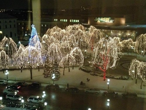 A snowy scene with trees adorned in colorful holiday lights, including a blue Christmas tree, in a city square at night.