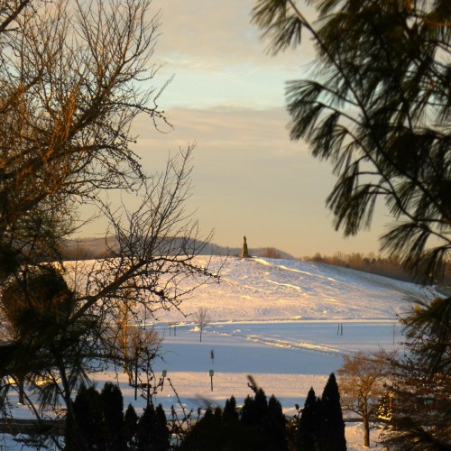A snowy landscape with distant hills and a tall monument, framed by trees in the foreground under a soft sunset sky.