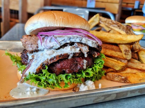 A juicy burger stacked with lettuce, onions, and sauce, served with a side of crispy fries on a metal tray.