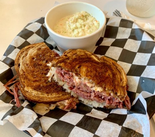 A toasted sandwich filled with shredded meat and sauerkraut, served with a bowl of coleslaw on a checkered tray.