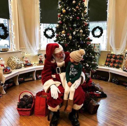 Santa Claus sits with a young child in front of a decorated Christmas tree, surrounded by festive decorations.