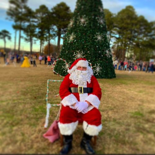 Santa Claus sits in front of a large Christmas tree, surrounded by festive decorations and a crowd in the background.