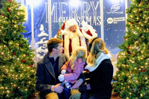 A family with a young girl sits in front of Santa Claus, surrounded by festive decorations and Christmas trees.