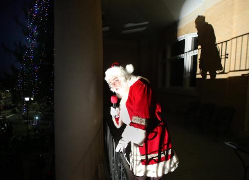Santa Claus in a red suit leans over a balcony, holding a microphone, with a shadow cast behind him.