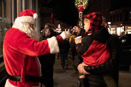 A man in a Santa costume interacts with a child held by an adult, festive lights in the background.