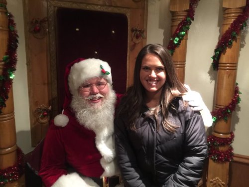 A smiling woman stands next to Santa Claus in a festive setting decorated with tinsel.