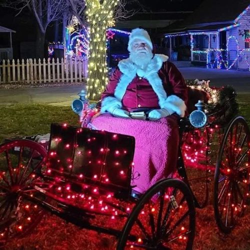 A man dressed as Santa sits in a decorated sleigh with colorful lights, surrounded by festive holiday decorations.