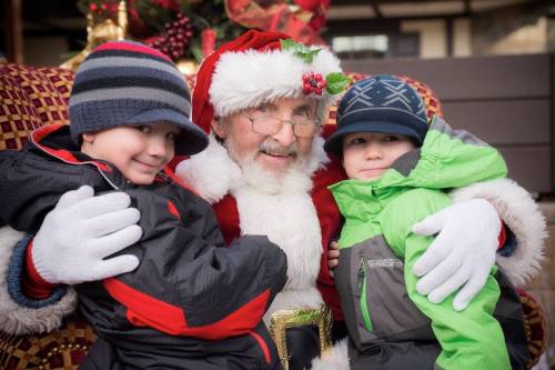 Santa Claus sits with two smiling children, one in a green jacket and the other in a black jacket, both wearing hats.