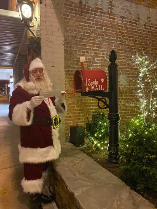 Santa Claus stands by a red mailbox labeled "Santa Mail," holding a letter in a festive, decorated street.