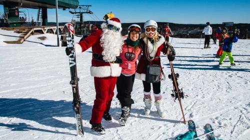 A Santa Claus figure poses with two women in ski gear on a snowy slope, with a ski lift in the background.