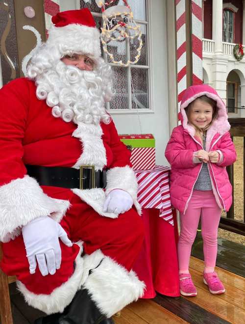 A smiling child in a pink coat stands next to Santa Claus, who is dressed in a red suit with a white beard.