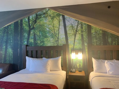 A hotel room with two beds, featuring a forest mural on the wall and warm lighting.