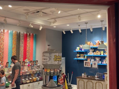 A colorful candy shop interior with a candy dispenser, shelves of treats, and a customer browsing.