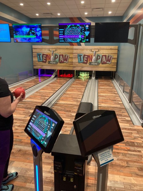 A person prepares to bowl at a modern bowling alley with screens and a wooden floor. Ten-pin setup visible.