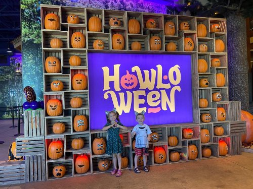 Two children pose in front of a colorful "Howl-O-Ween" display with carved pumpkins and a festive backdrop.