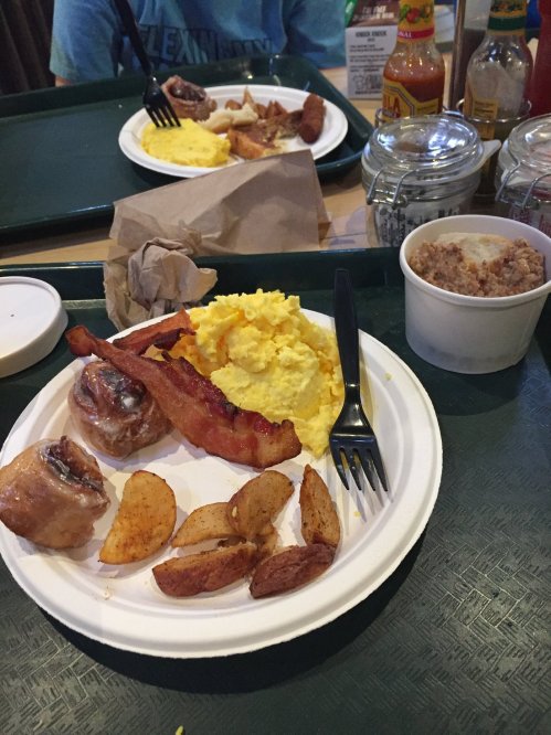 A plate of breakfast with scrambled eggs, bacon, potatoes, and cinnamon rolls, served on a tray.
