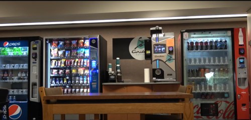 A vending area featuring snack and beverage machines, with a table in the foreground and a logo on the wall.