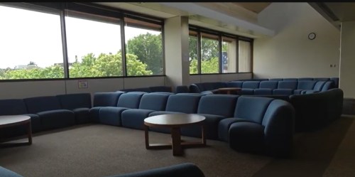 A spacious lounge area with blue couches and wooden tables, featuring large windows and natural light.