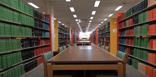 A spacious library with rows of colorful bookshelves and a central table surrounded by chairs.
