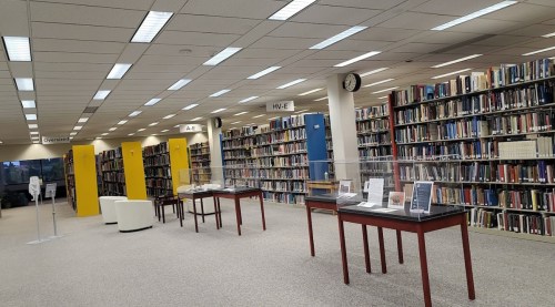 A spacious library interior with bookshelves, display tables, and seating areas under bright overhead lights.