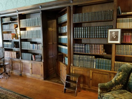 A wooden bookshelf filled with books, featuring a hidden door and a framed photo on the wall beside a cozy armchair.