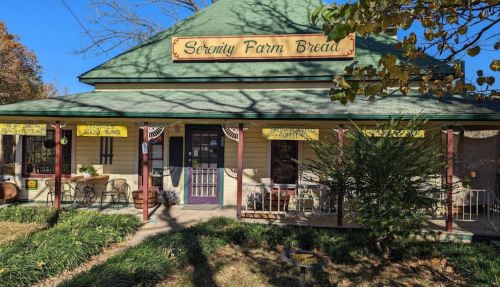 A quaint building with a green roof and sign reading "Serenity Farm Bread," surrounded by greenery and outdoor seating.