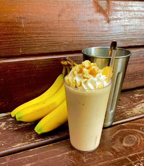 A creamy banana milkshake topped with whipped cream and cereal, next to fresh bananas and a metal cup on a wooden surface.