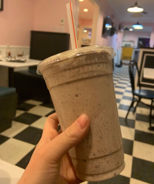 A hand holding a large cup of chocolate milkshake with a straw, set in a retro-style diner.