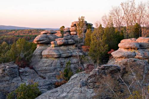 Rock formations surrounded by trees at sunset, showcasing layered stones and a colorful sky in the background.