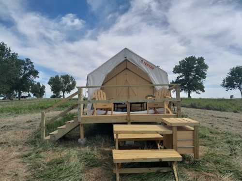 A glamping tent on a wooden deck, surrounded by grass and trees, with seating and a picnic table in front.