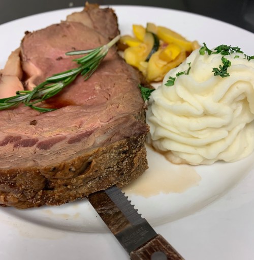 A plate featuring a slice of roast beef with rosemary, creamy mashed potatoes, and sautéed vegetables.