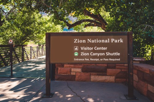 Sign for Zion National Park indicating the Visitor Center and Zion Canyon Shuttle, surrounded by greenery.