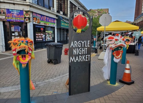 Colorful dragon masks flank a sign for Argyle Night Market, with shops and tents in the background.